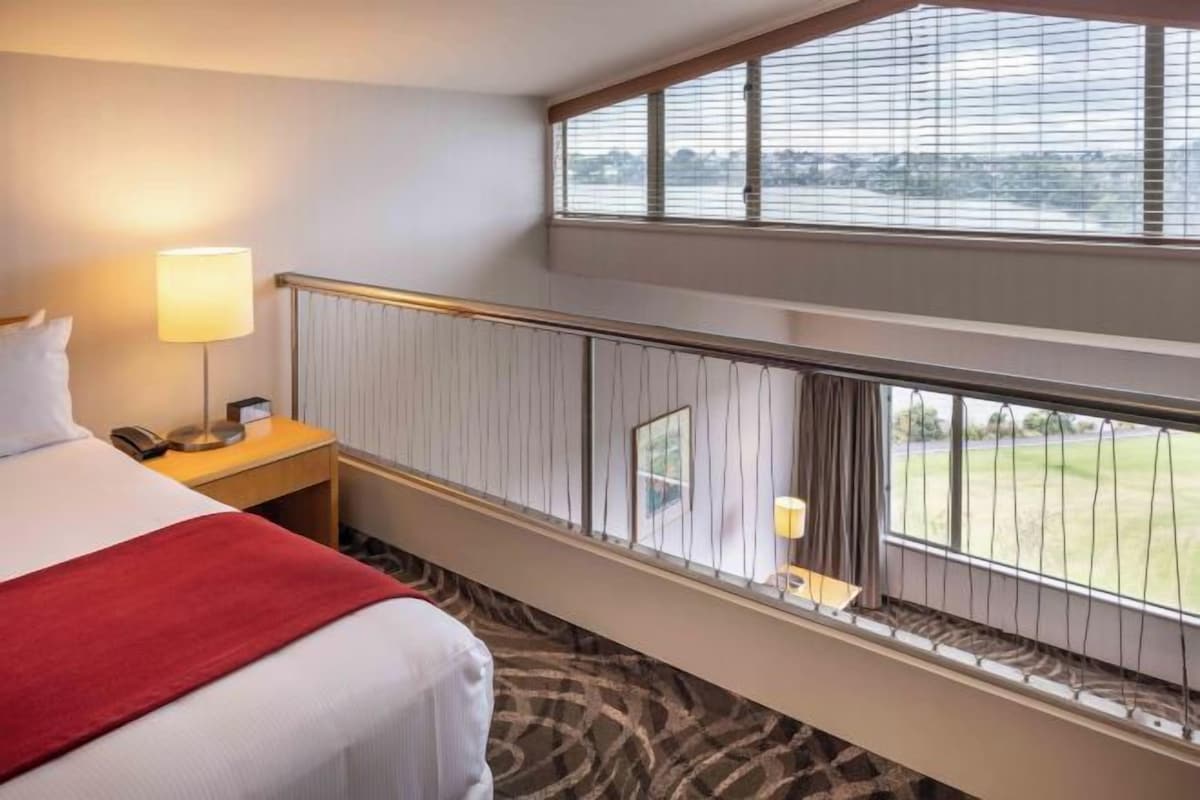 A hotel room with a bed, a nightstand, a lamp, a window with a view of a field, and a balcony railing.
