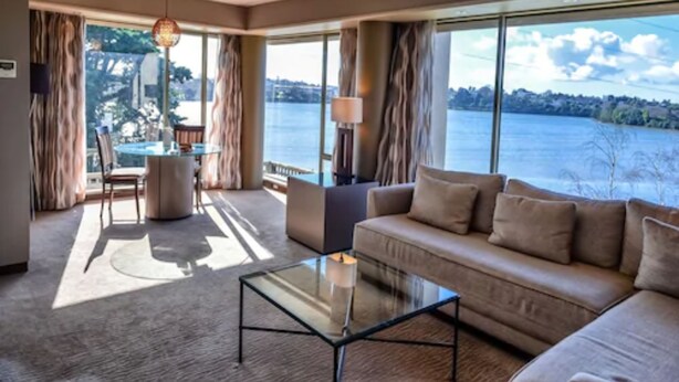 A spacious living room with a large window offering a view of a lake and trees. There is a sofa, a glass coffee table, and a dining area with a table and chairs. - Waipuna Hotel & Conference Centre (Auckland)