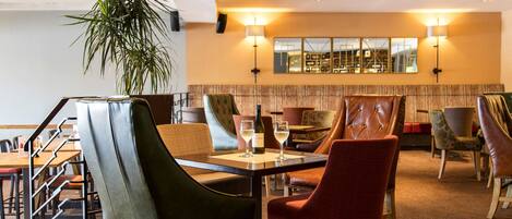 A hotel lobby with a dining area featuring leather chairs, a potted plant, and a large mirror on the wall.