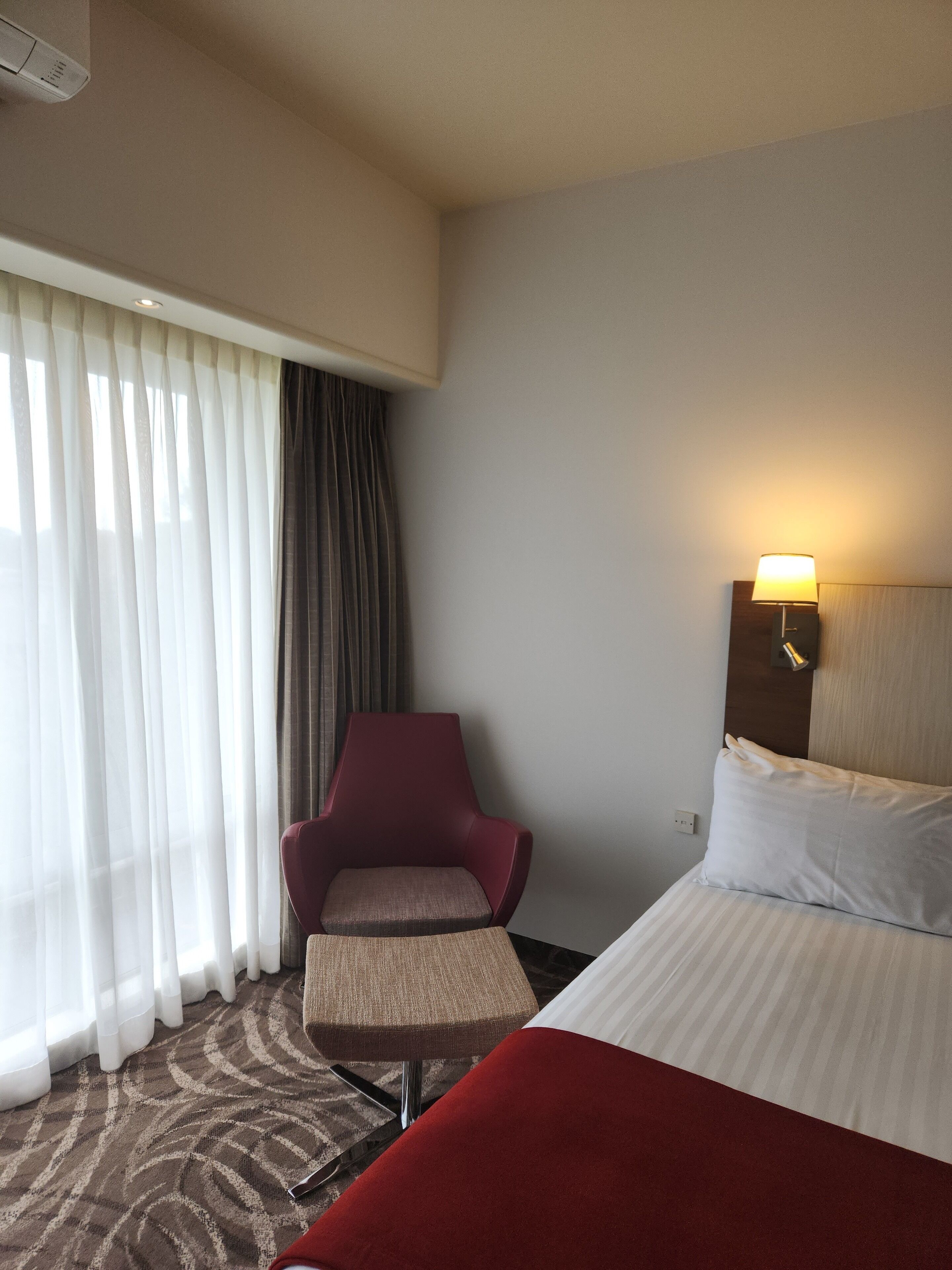 Une chambre d’hôtel avec un lit, un fauteuil rouge, une petite table, une lampe et une fenêtre avec des rideaux.