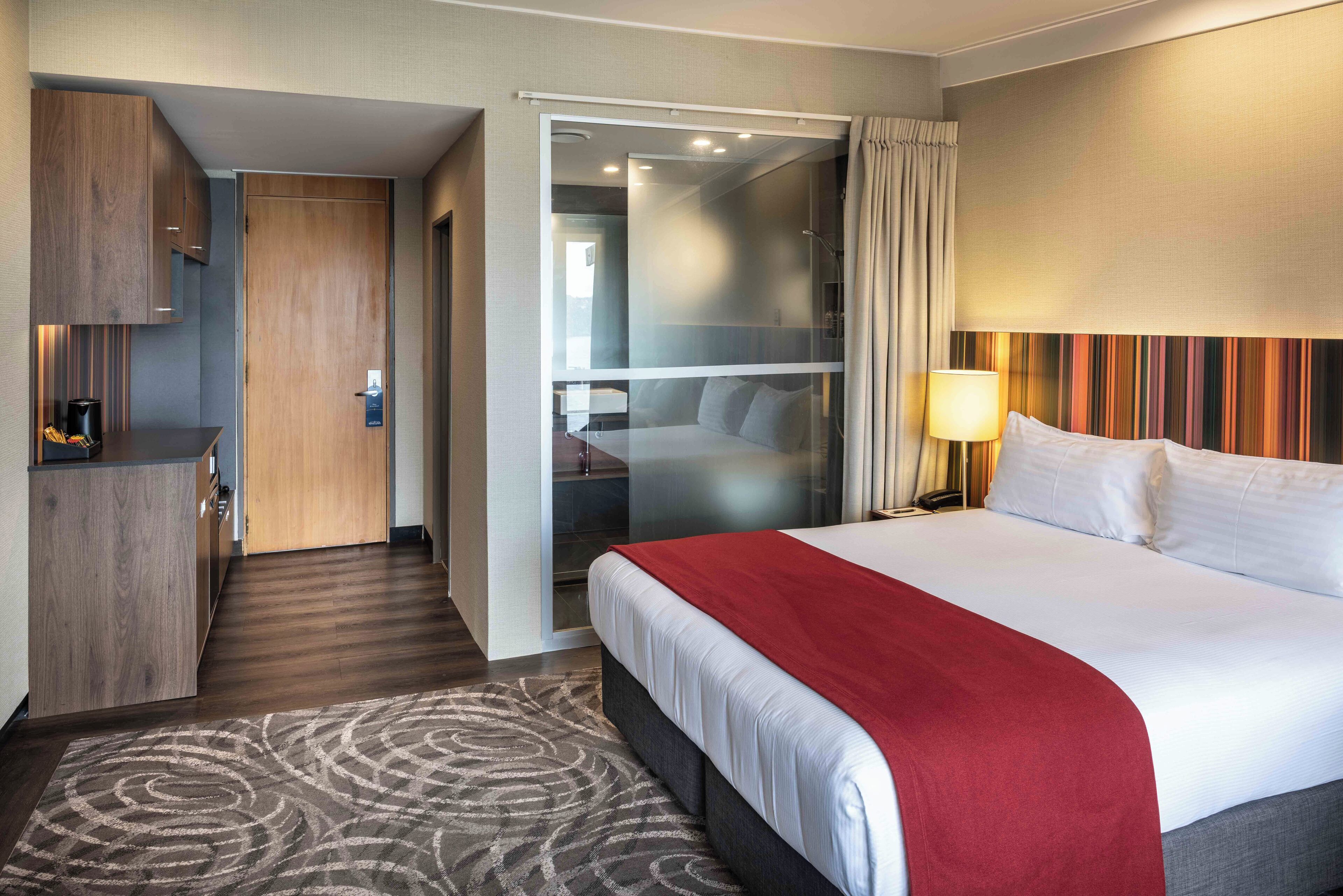 A hotel room with a large bed, a desk, a mirror, and a sliding door leading to a balcony.