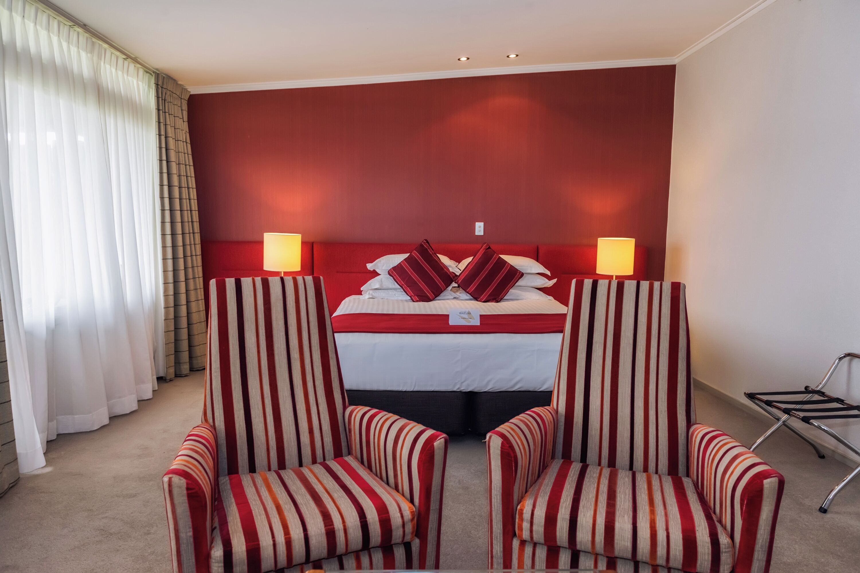 Ein Hotelzimmer mit einem rot gestreiften Kopfteil, zwei gestreiften Sesseln, einem Bett mit rot-weißer Bettwäsche und einem Nachttisch mit einer Lampe.
