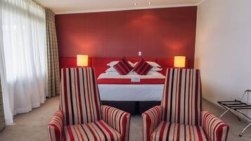 Ein Hotelzimmer mit einem rot gestreiften Kopfteil, zwei gestreiften Sesseln, einem Bett mit rot-weißer Bettwäsche und einem Nachttisch mit einer Lampe.
