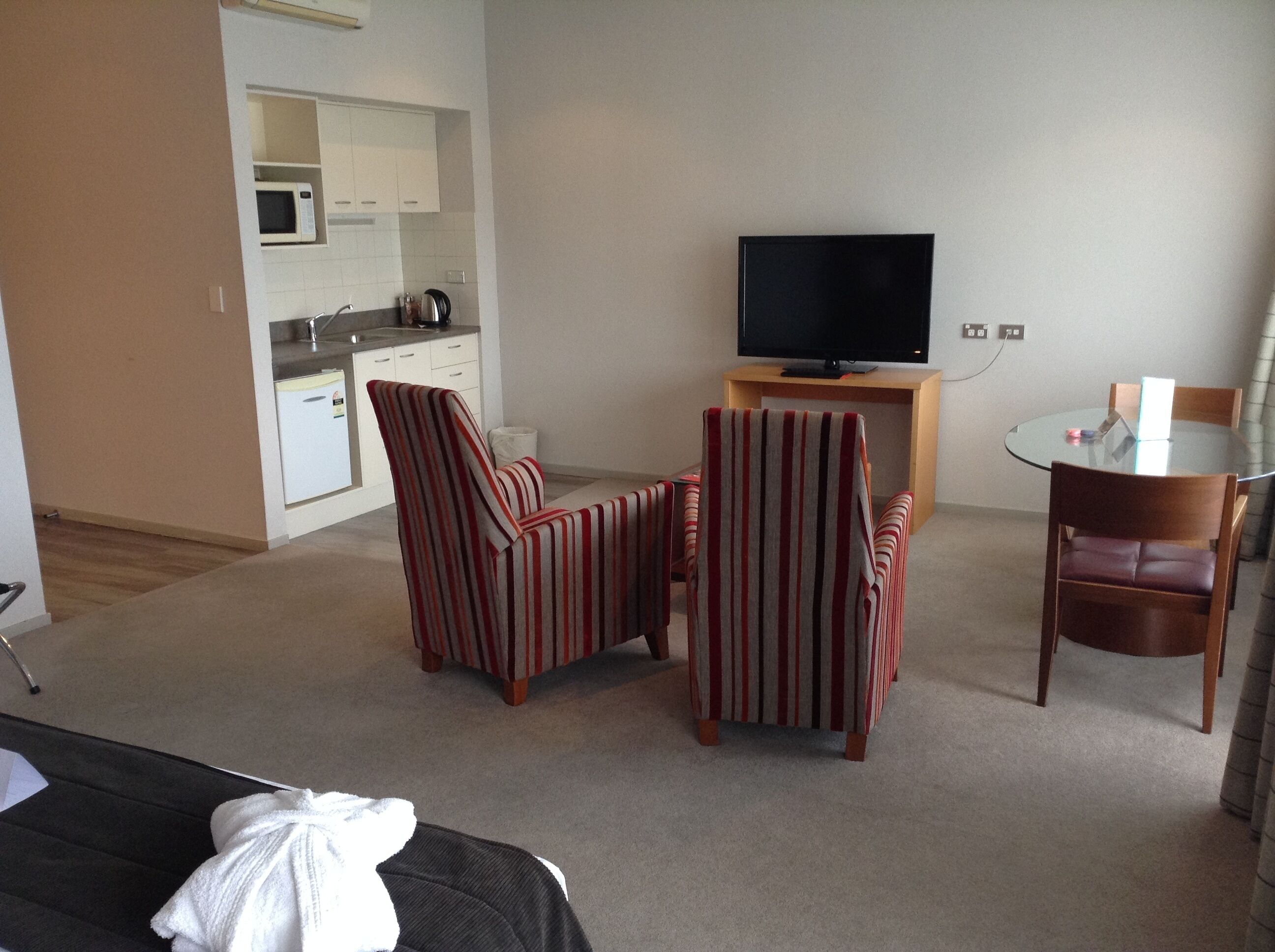 A compact hotel room with a kitchenette, a TV on a wooden stand, striped armchairs, a small round table, and a bed with a white blanket.