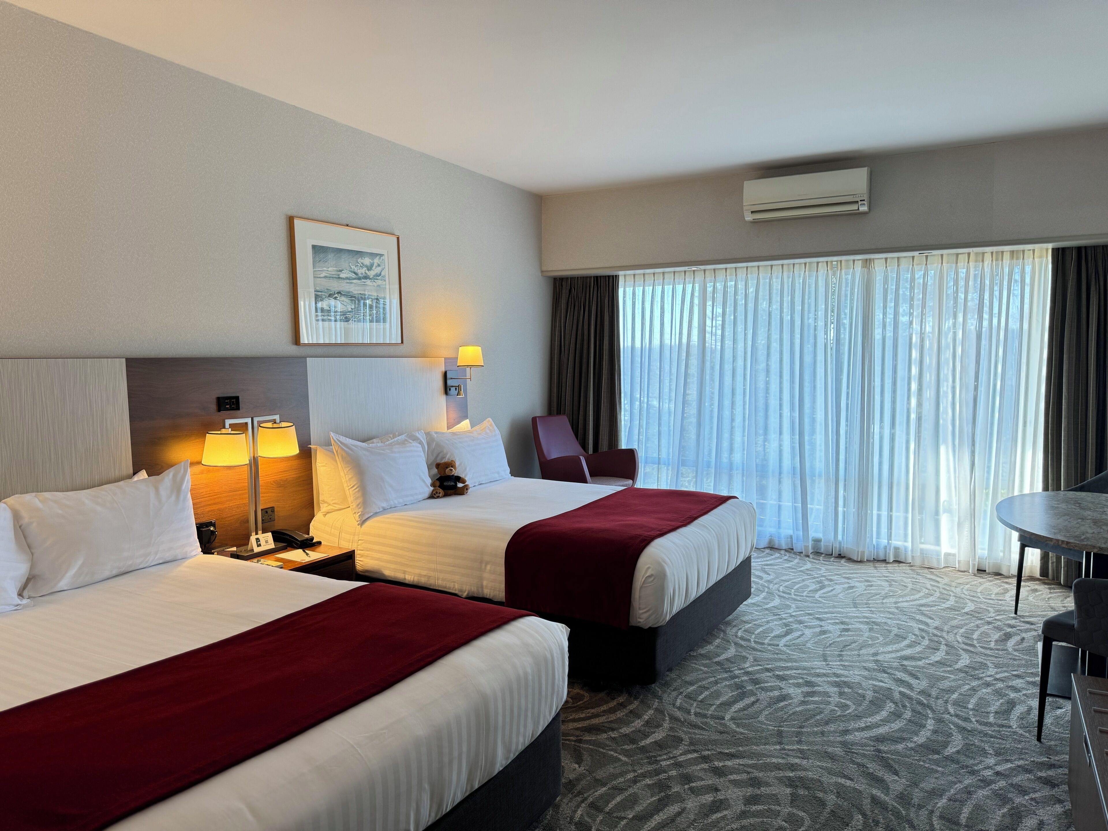 Ein Hotelzimmer mit zwei Betten, einem Schreibtisch und einem großen Fenster mit Vorhängen.