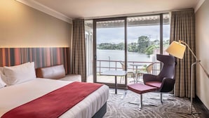 A hotel room with a large bed, a sofa, a small table, a chair, and a view of a body of water through large windows. - Waipuna Hotel & Conference Centre (Auckland)