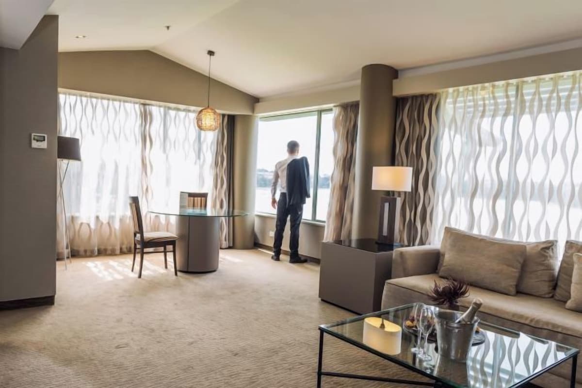 A hotel room with a sofa, a glass coffee table, a chair, a lamp, and a view of the outdoors.