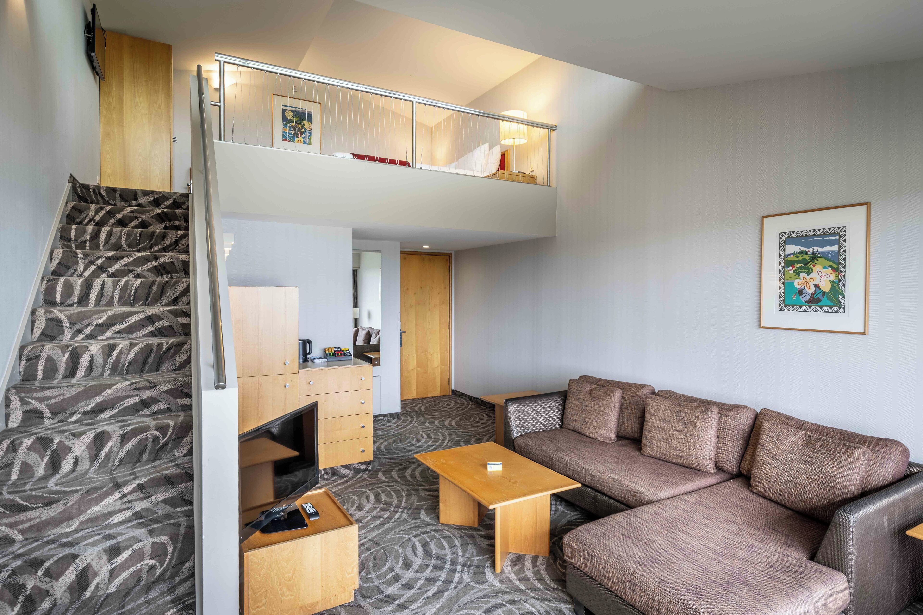 A modern hotel room with a staircase leading to a loft bedroom, a sofa, a small table, and a wooden cabinet.