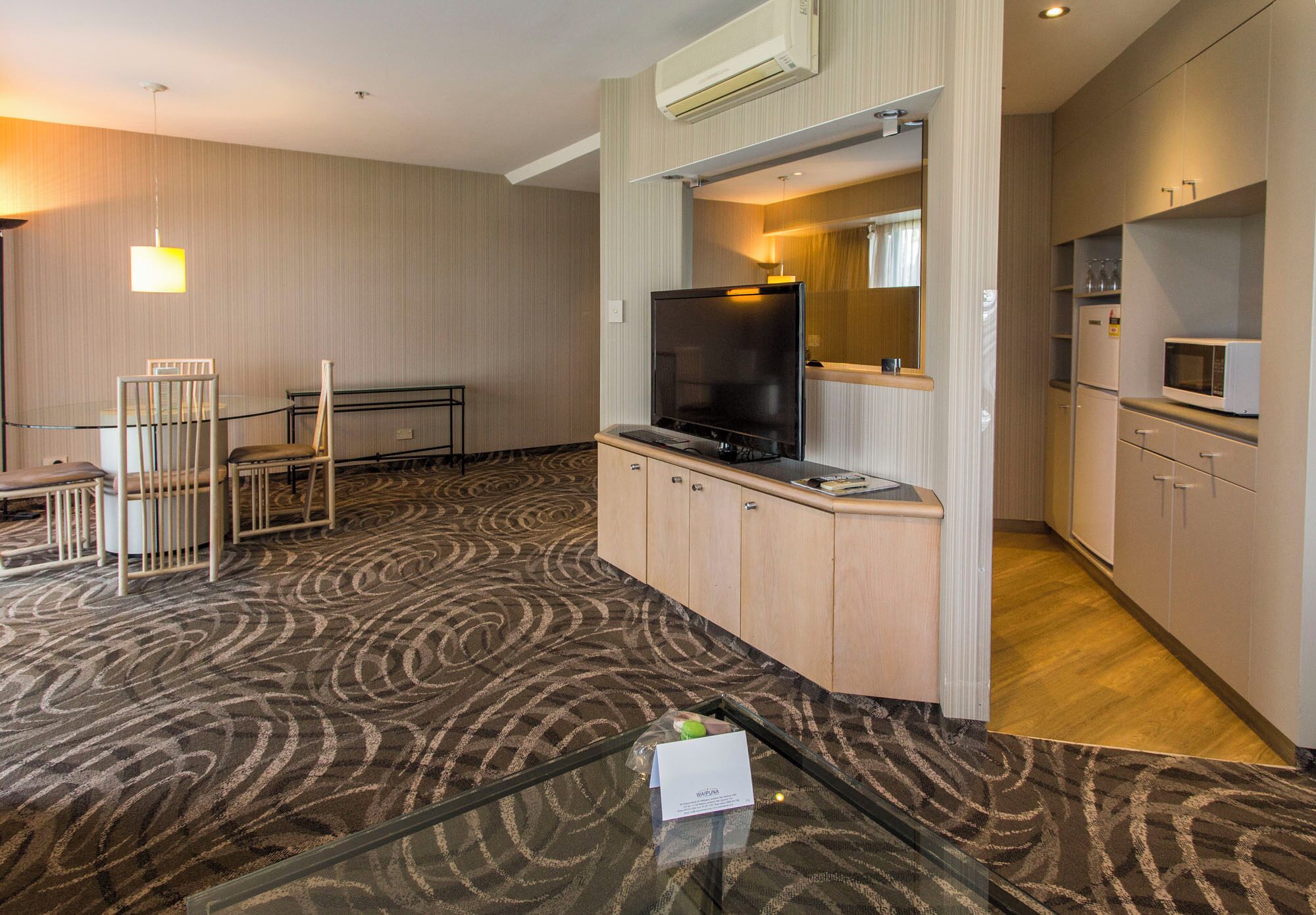 A hotel room with a TV, kitchenette, and a patterned carpet.