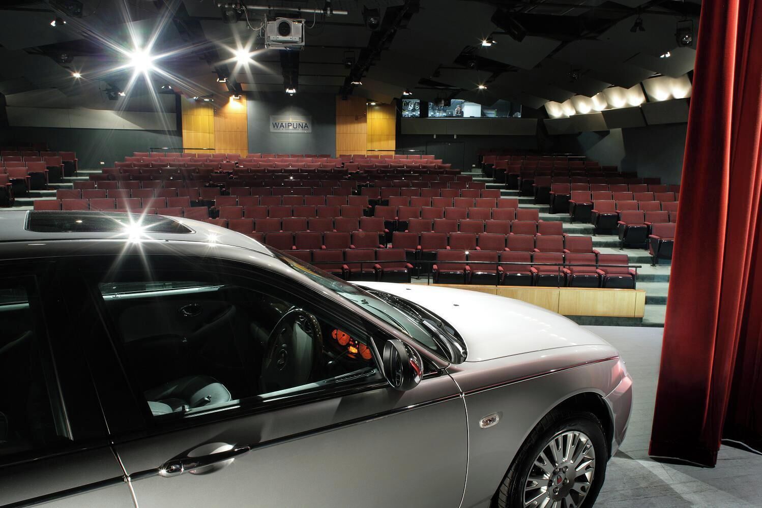 A car parked in a modern indoor setting with rows of red seats and a stage area labeled "WAIPUNA".