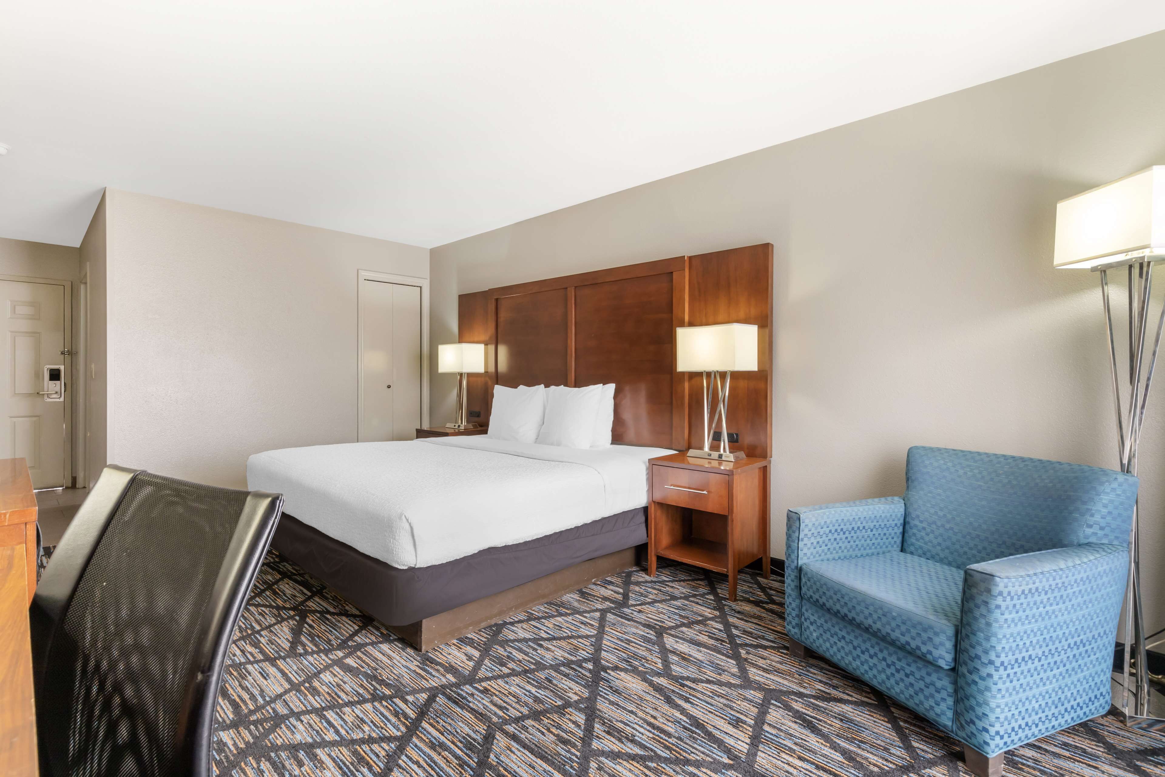 A hotel room with a bed, bedside tables with lamps, a blue armchair, and a patterned carpet.
