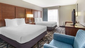 A hotel room with two beds, a desk with a flat-screen TV, a blue armchair, and a large window with curtains. - Best Western near Lackland AFB/SeaWorld (San Antonio)