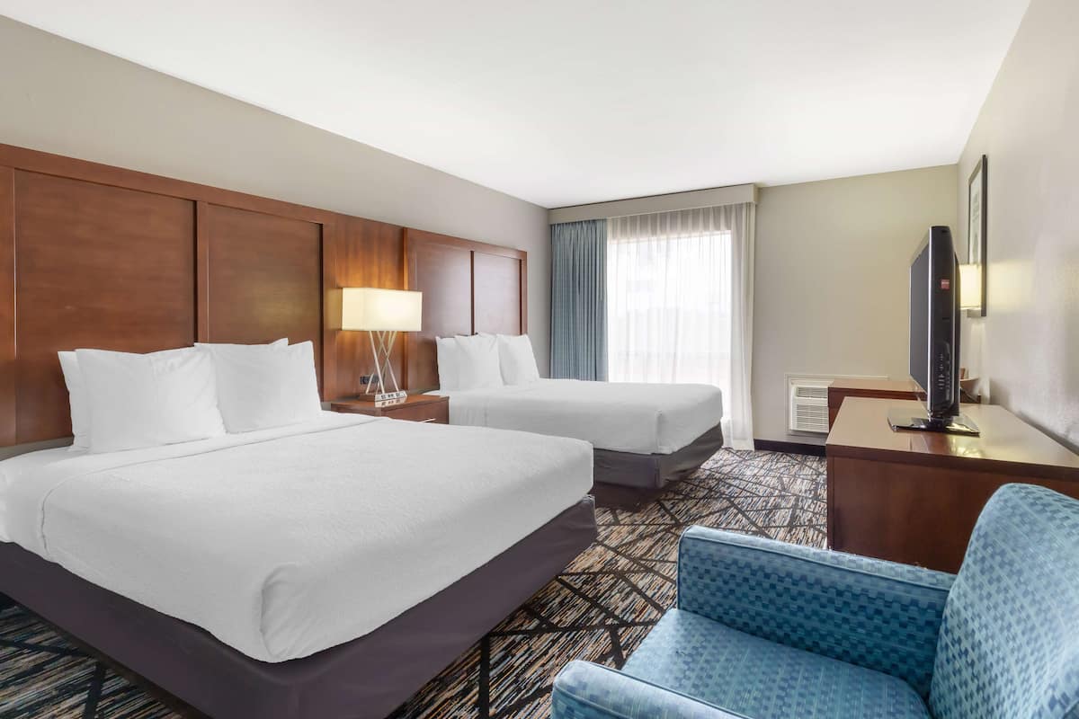 A hotel room with two beds, a desk with a flat-screen TV, a blue armchair, and a large window with curtains.
