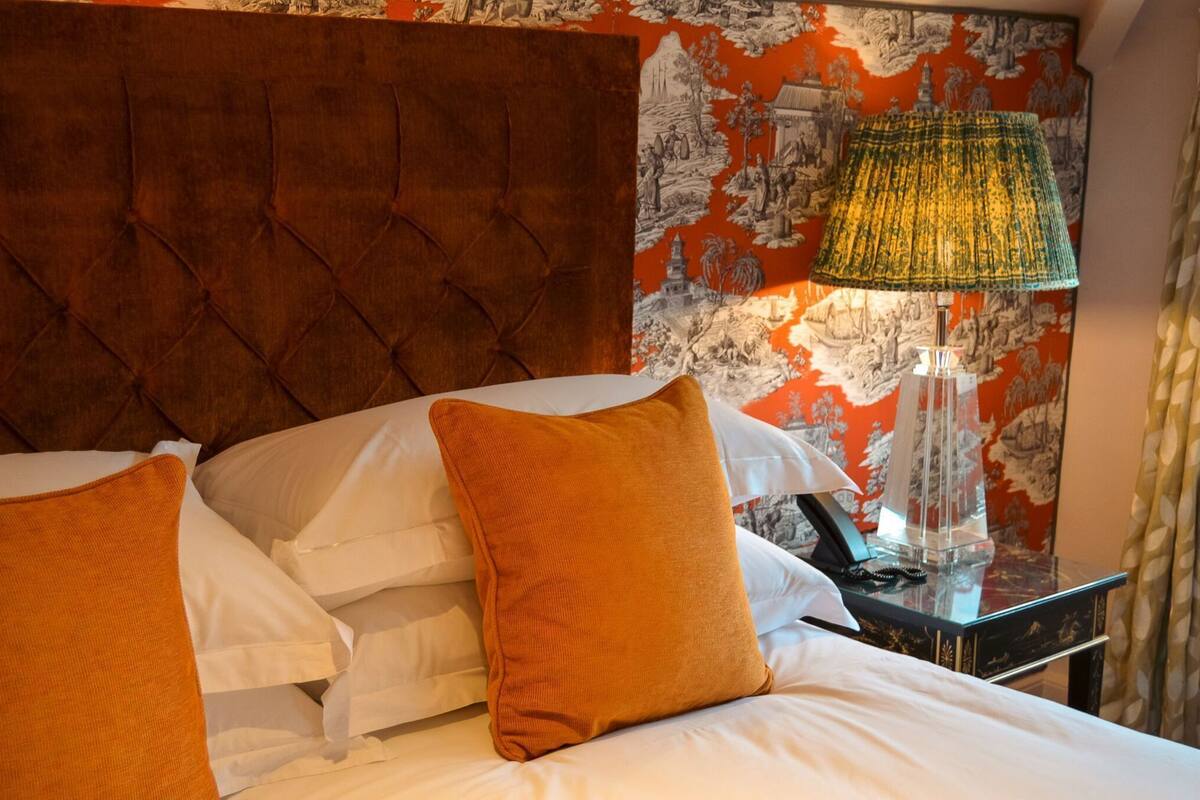 A hotel room with a tufted headboard, a bed with white and orange pillows, a nightstand with a lamp, and a wall with a red and black patterned wallpaper.