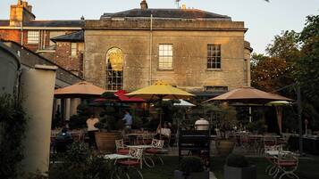 An outdoor patio area with tables and chairs, surrounded by a brick building with arched windows and a clock tower.