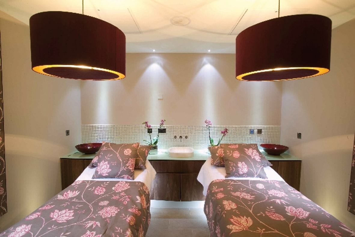 A room with two beds, a sink, and a mirror. There are two large lamps hanging from the ceiling.