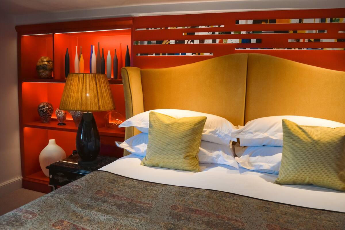 A hotel room with a bed, a nightstand with a lamp, and a red bookshelf with decorative items.