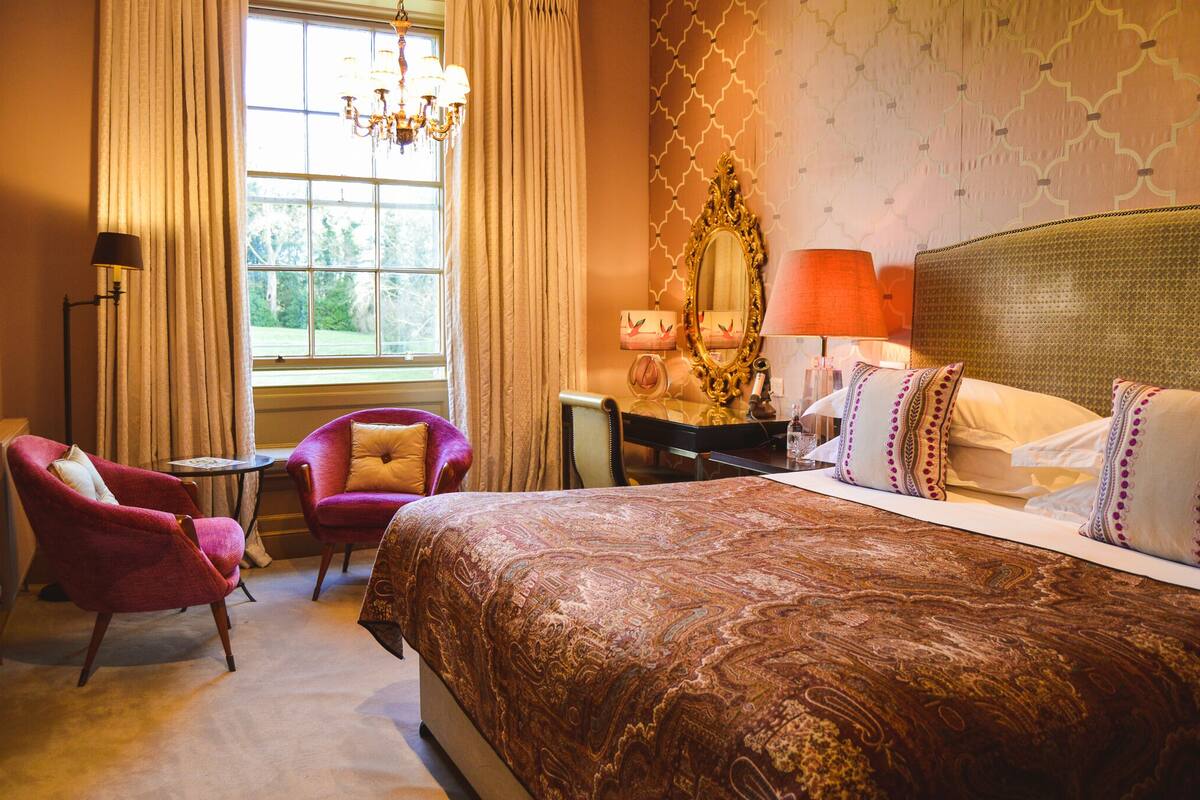 A hotel room with a large bed, two armchairs, a desk, a chandelier, and a window with curtains.