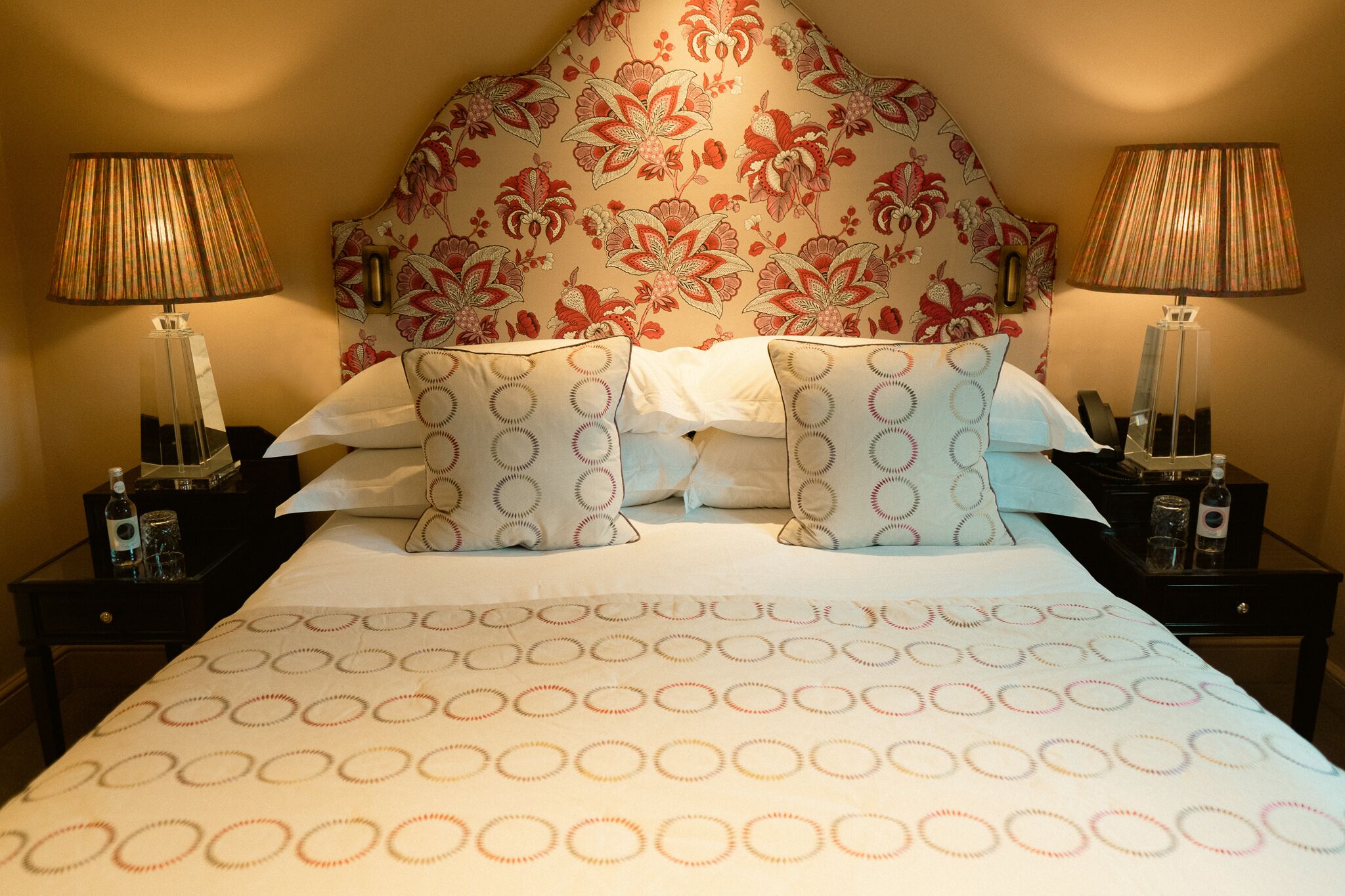 Habitación de hotel con un cabecero floral, dos mesitas de noche con lámparas, una cama con ropa de cama estampada y un buró con una botella y vasos.