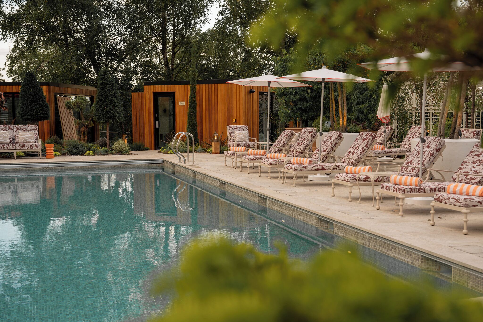Ein Poolbereich mit Liegestühlen und Sonnenschirmen, umgeben von Grünflächen und einem Holzgebäude.