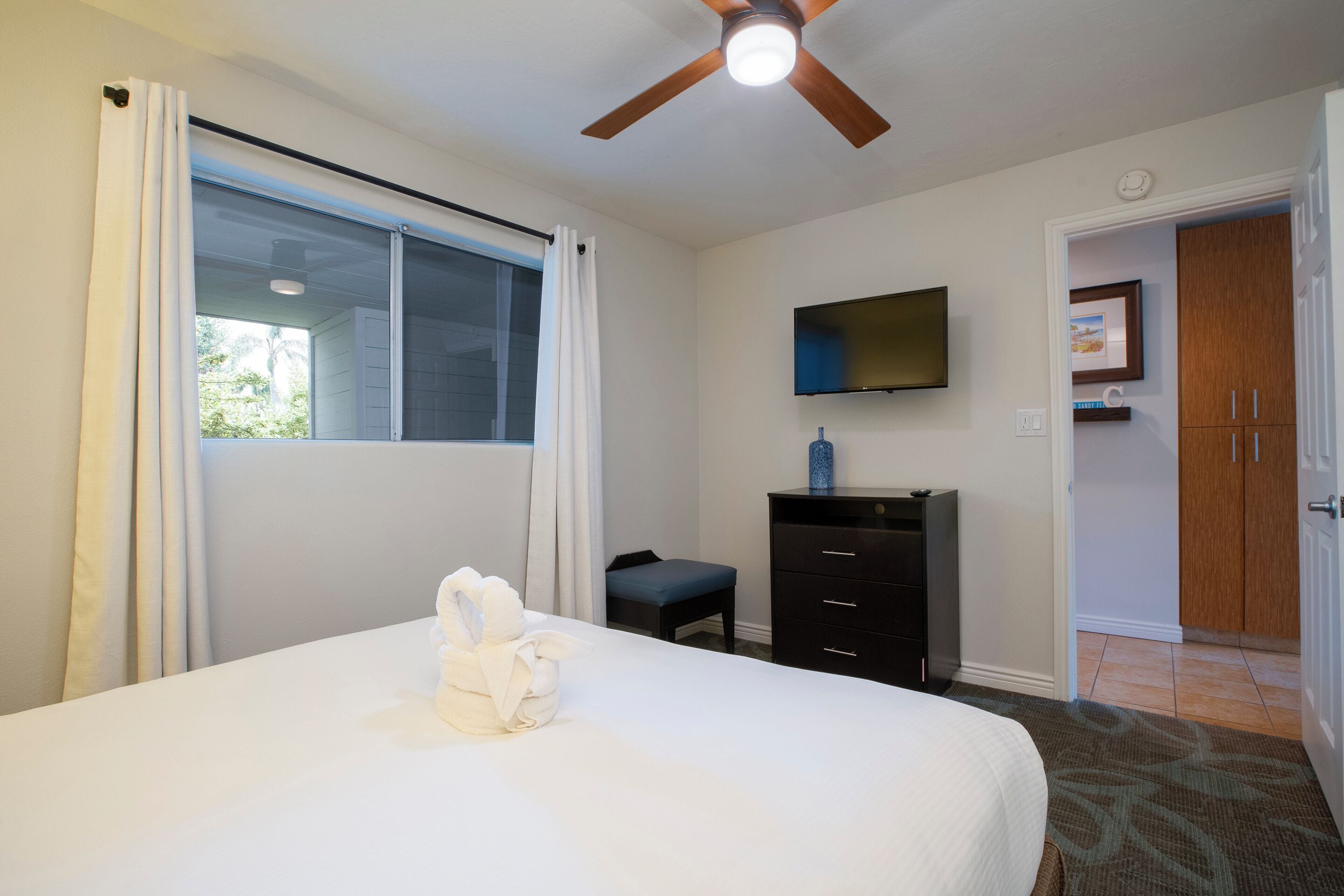 A hotel room with a bed, a ceiling fan, a TV mounted on the wall, a wooden wardrobe, and a window with curtains.