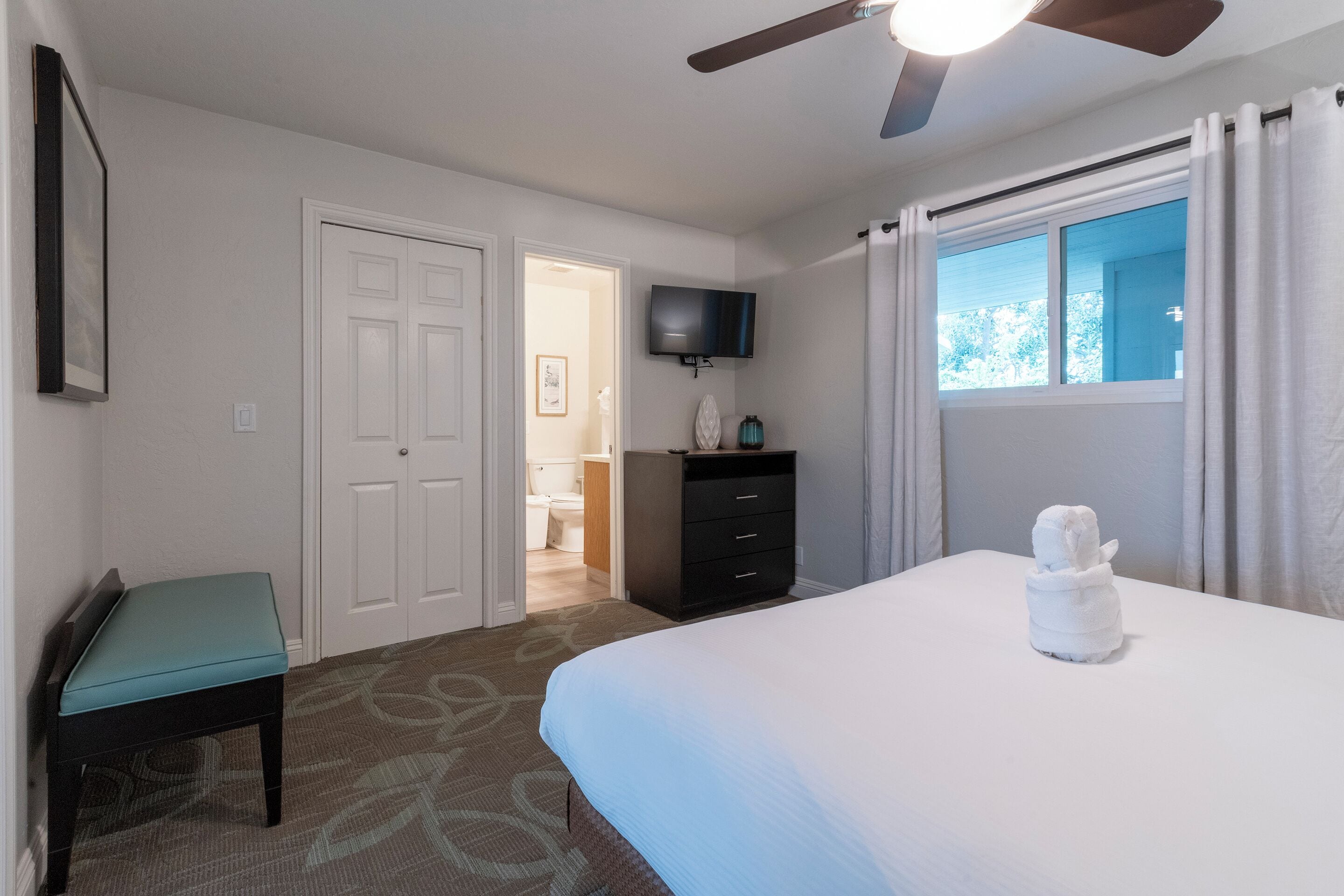 A hotel room with a bed, a desk, a chair, a television, a ceiling fan, and a window with curtains.