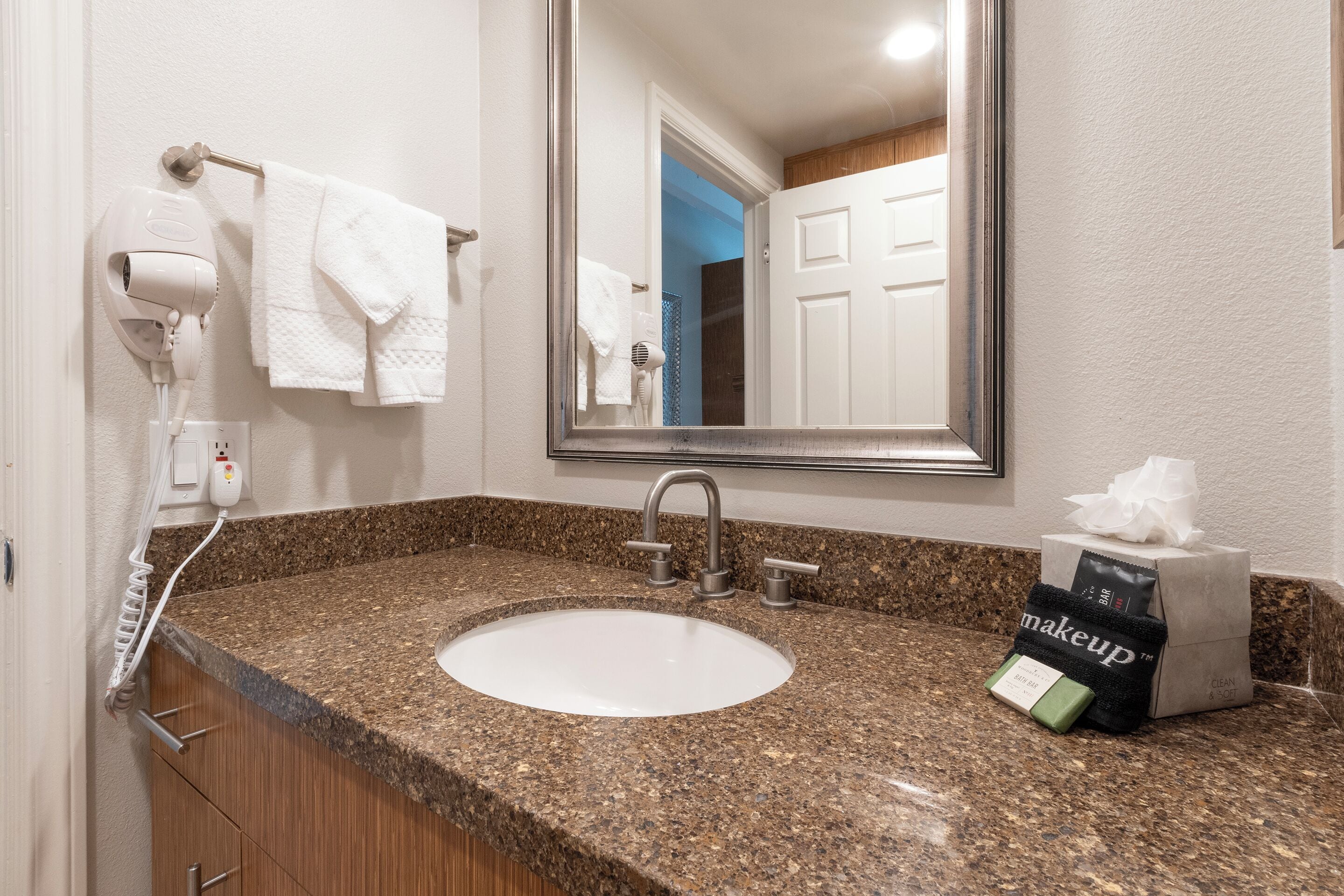 Une salle de bain dotée d’un comptoir en granit, d’un lavabo blanc, d’un miroir, d’un porte-serviettes, d’un sèche-cheveux et d’une trousse de maquillage.