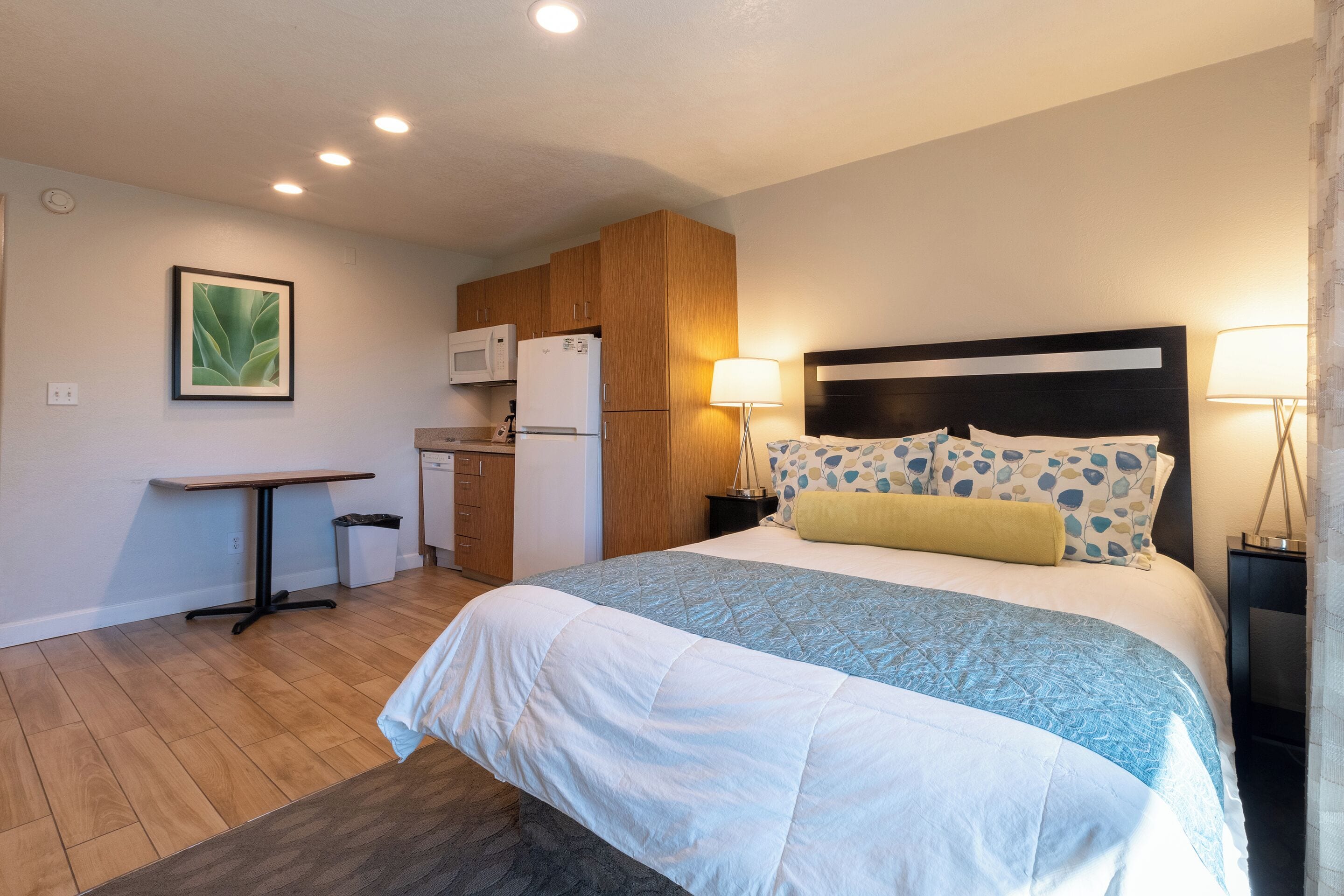 A bedroom with a bed, bedside lamps, a nightstand, a wooden headboard, a small table, and a kitchenette with a microwave and refrigerator.