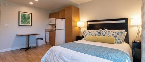 A bedroom with a bed, bedside lamps, a nightstand, a wooden headboard, a small table, and a kitchenette with a microwave and refrigerator.