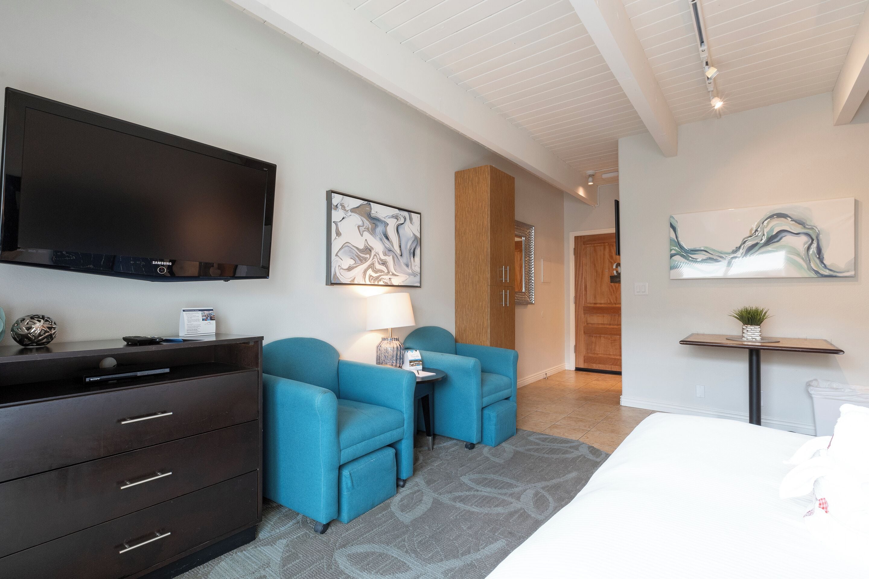 A modern hotel room with a flat-screen TV, a wooden cabinet, two blue armchairs, a small table with a lamp, and abstract wall art.