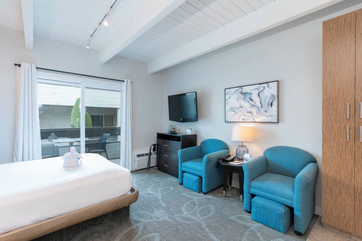 A hotel room with a bed, two blue armchairs, a TV, a desk, and a wooden wardrobe.