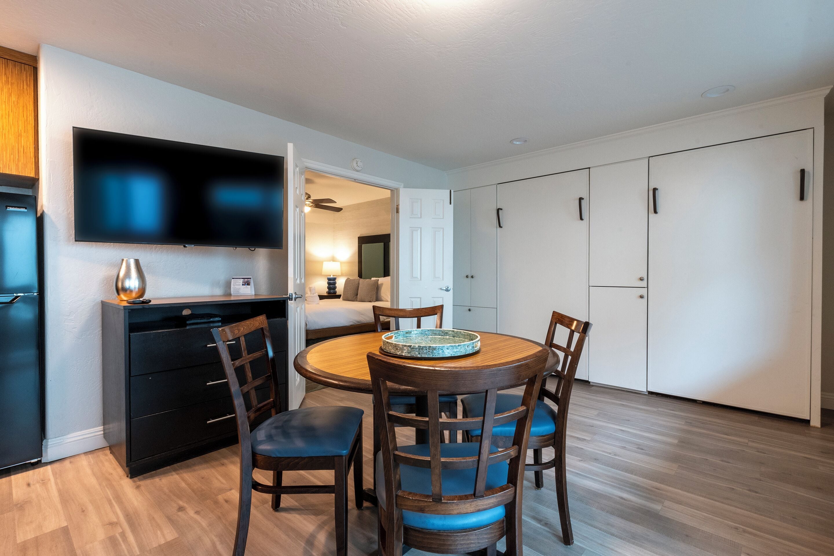 A modern kitchen with a dining area, a TV mounted on the wall, and a ceiling fan.