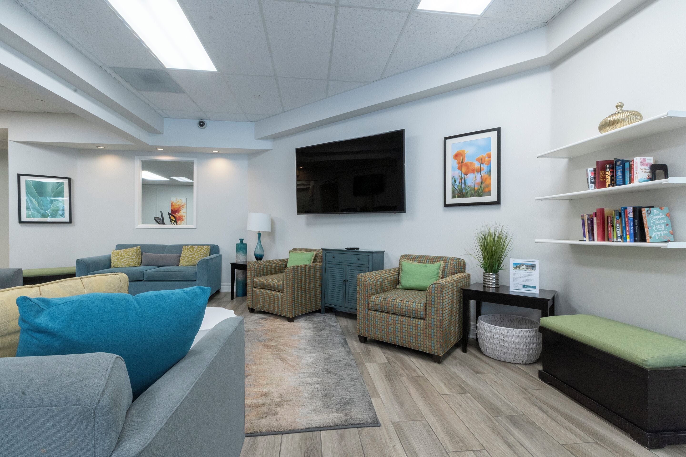 A modern living room with a grey sofa, blue armchair, and a wicker armchair with green cushions. A flat-screen TV is mounted on the wall, and there are bookshelves with books and decorative items.