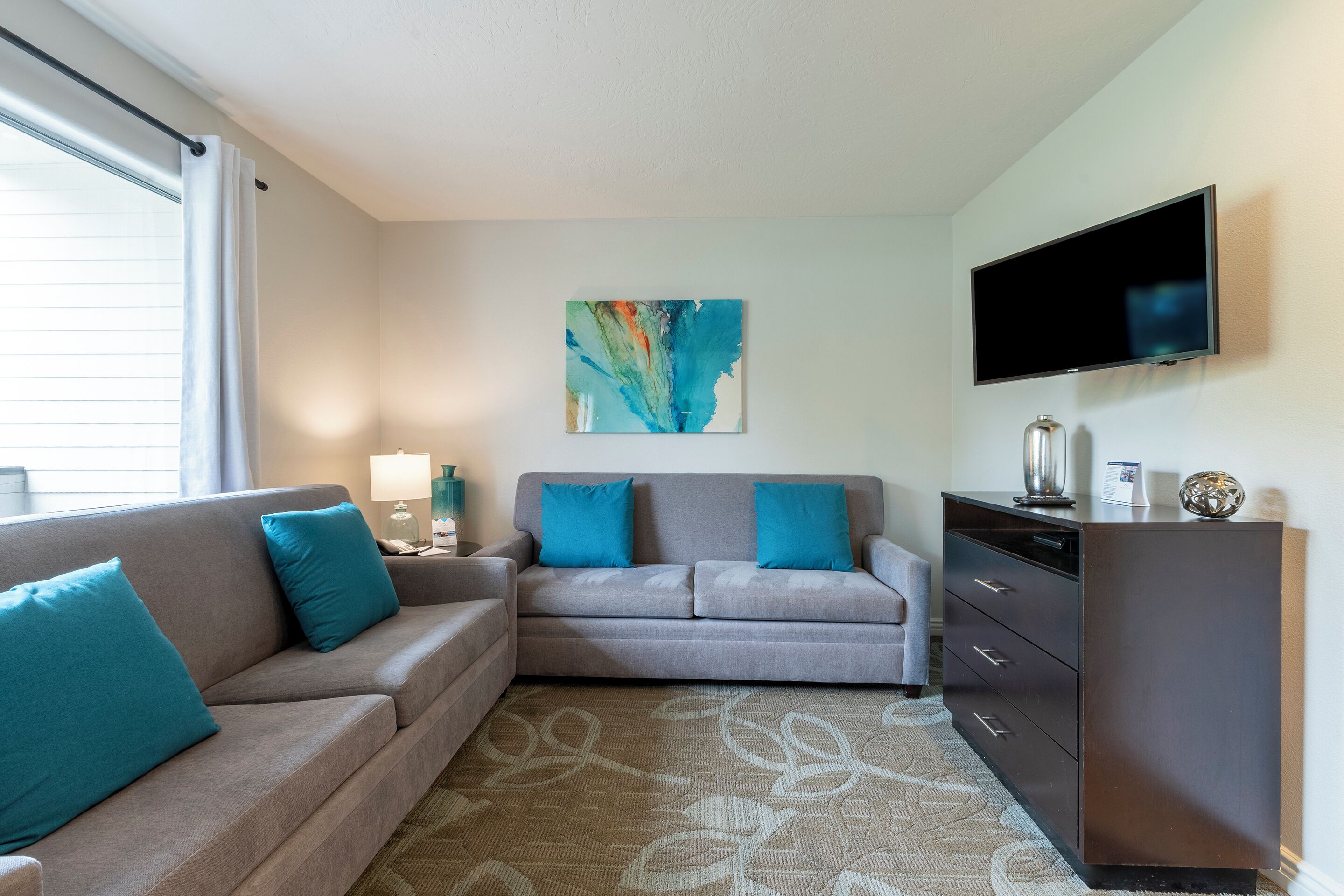 A modern living room with a grey sofa, teal cushions, a flat-screen TV, and a decorative abstract painting on the wall.