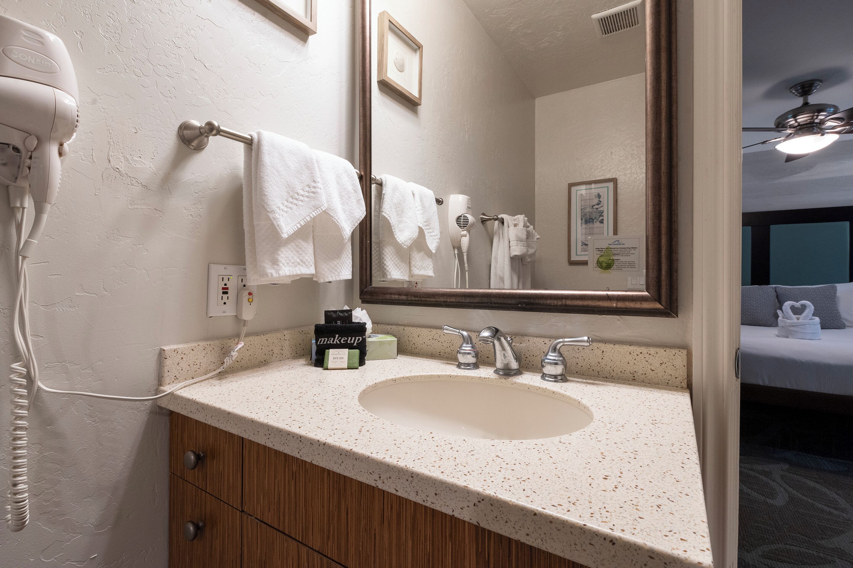 A hotel bathroom with a sink, towel rack, hair dryer, and a bed in the background.