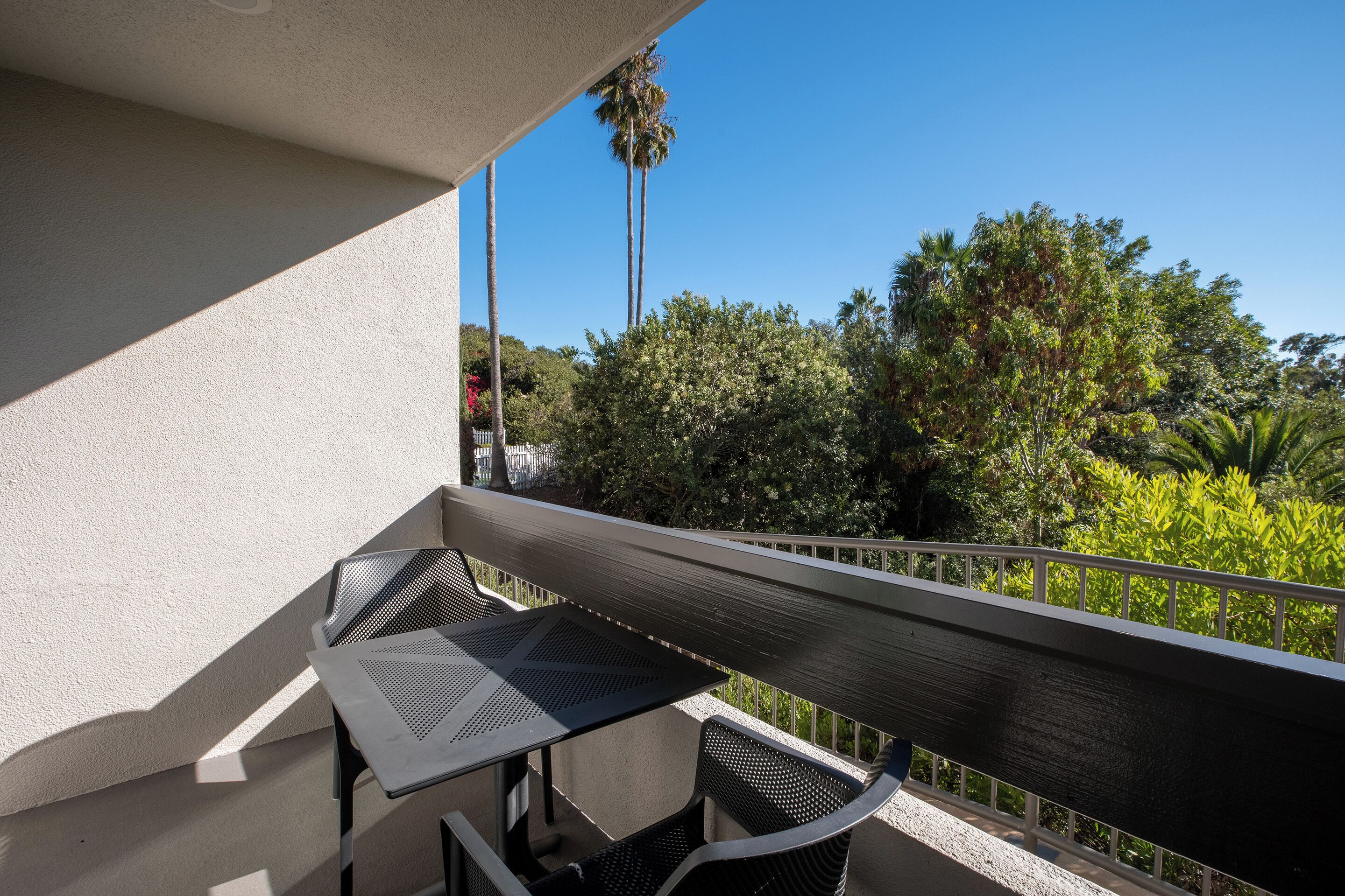 A balcony with a table and chairs, overlooking a garden with trees and a clear blue sky.