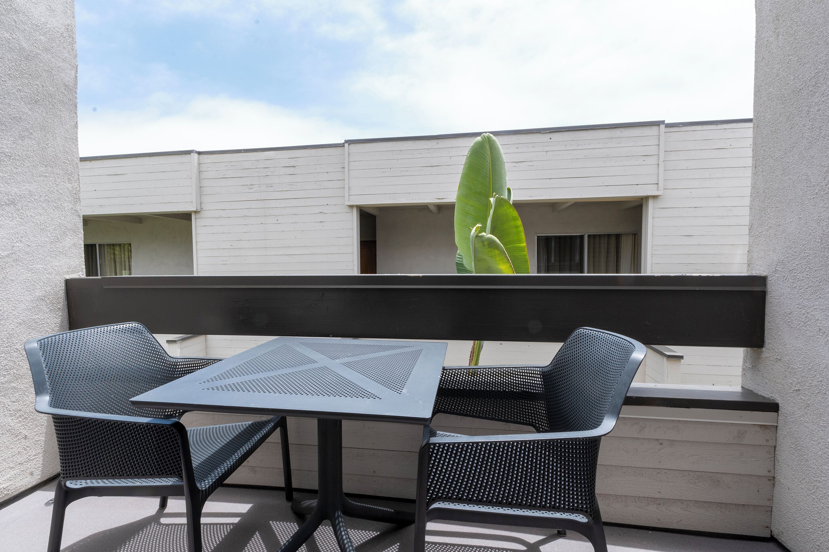 Un balcón con una mesa y dos sillas, una planta en maceta y un edificio al fondo.