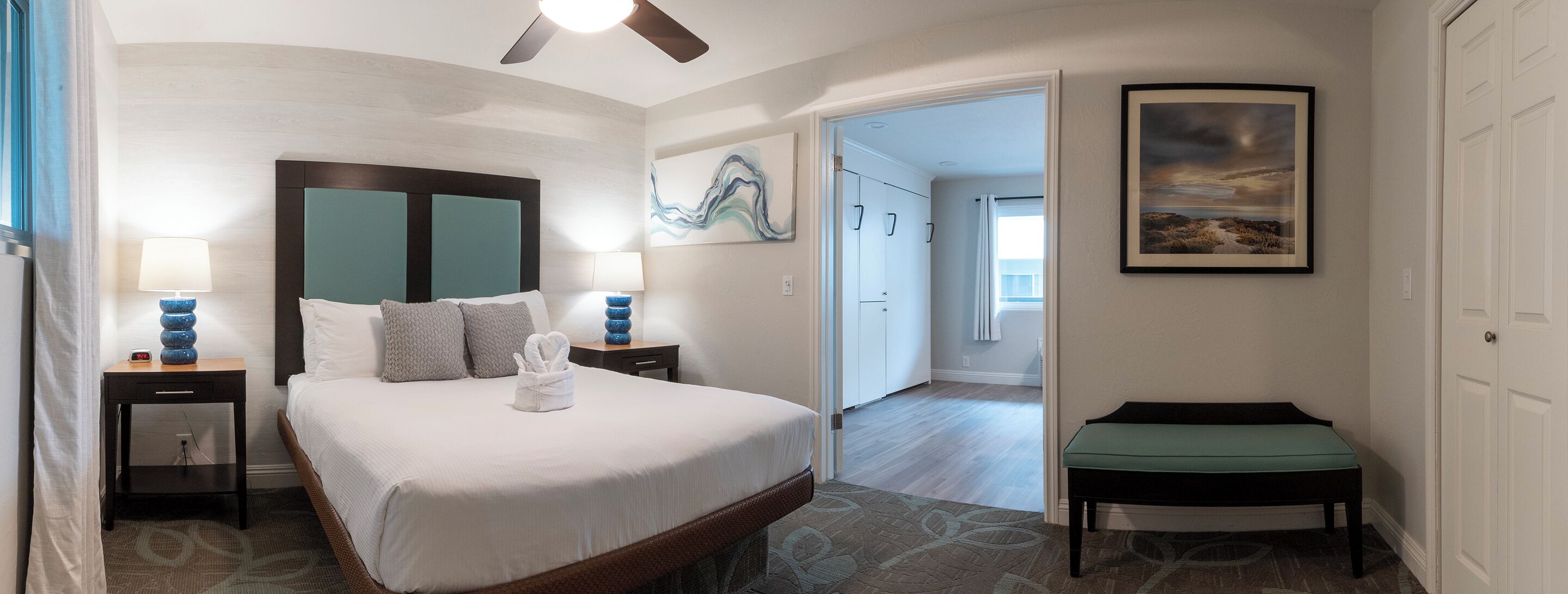 A hotel room with a large bed, two bedside tables with lamps, a bench, a painting, and a view of the hallway.