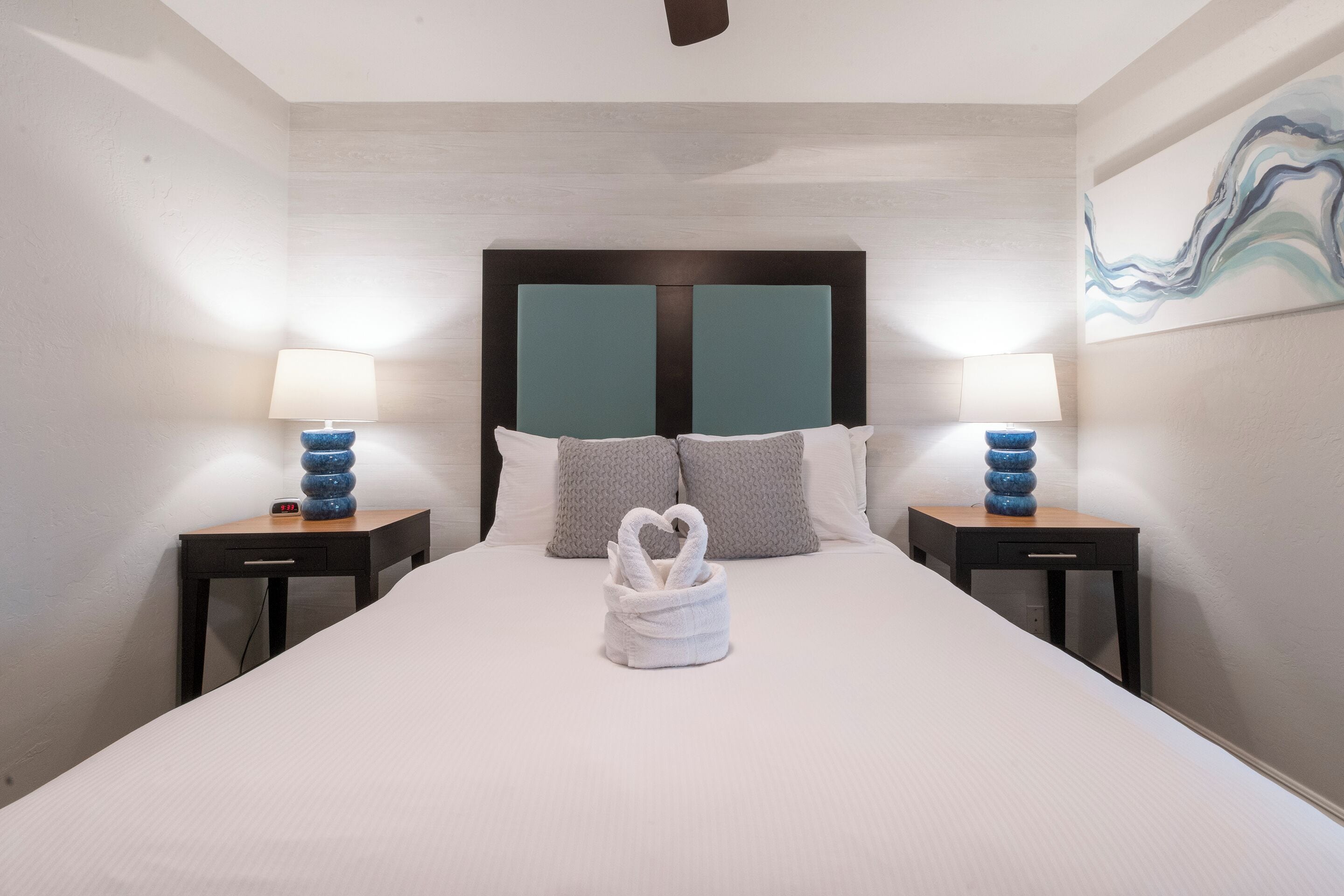 A neatly made bed with a heart-shaped towel, two bedside tables with lamps, and a painting on the wall.