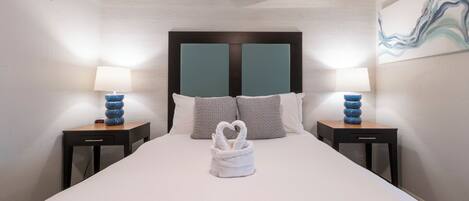 A neatly made bed with a heart-shaped towel, two bedside tables with lamps, and a painting on the wall.