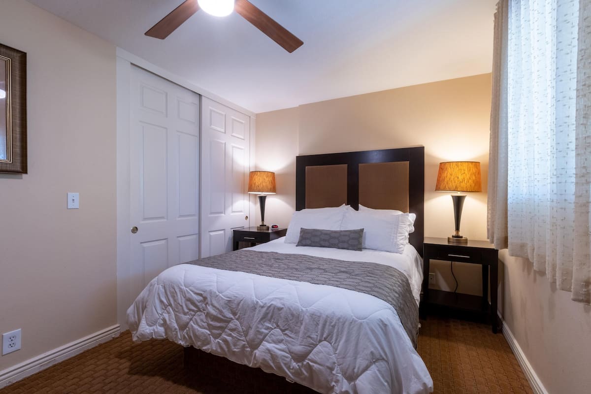 A bedroom with a bed, two bedside tables with lamps, a ceiling fan, a door, and a window with curtains.