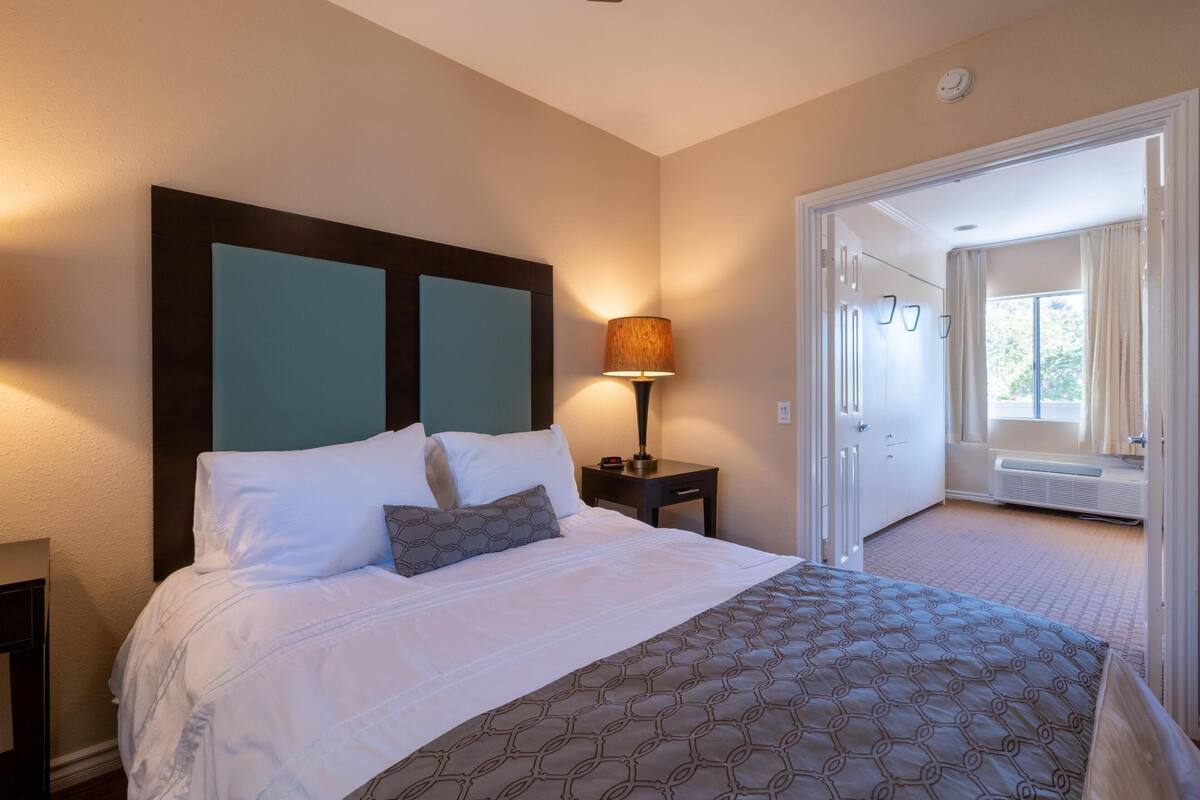 A hotel room with a bed, a nightstand, a lamp, a window, and an open door leading to another room.