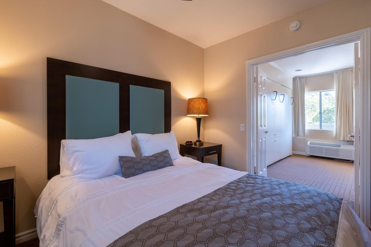 A hotel room with a bed, a nightstand, a lamp, a window, and an open door leading to another room.