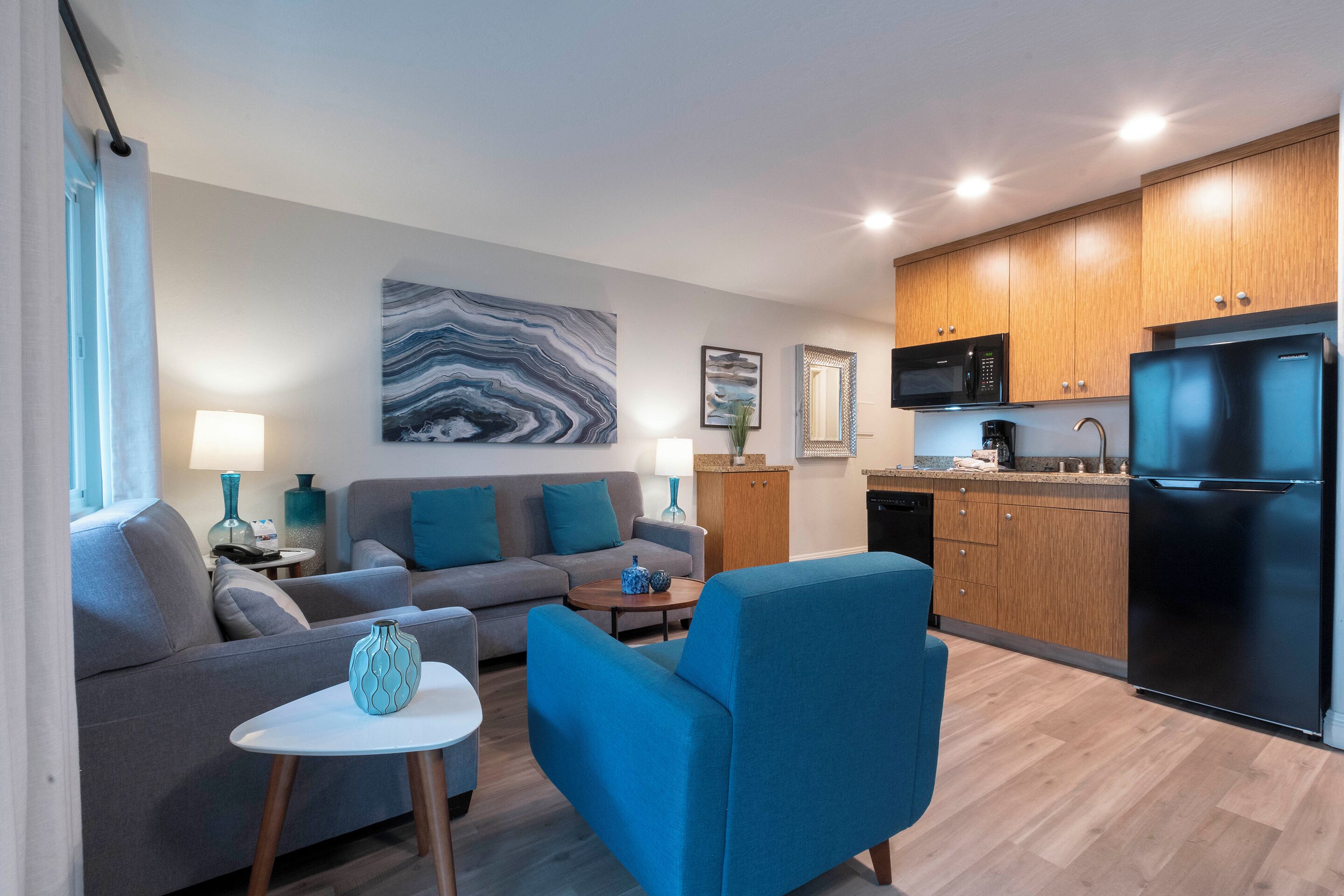 A modern living room with a grey sofa, a blue armchair, and a coffee table. There is a kitchen area with wooden cabinets and a black refrigerator.