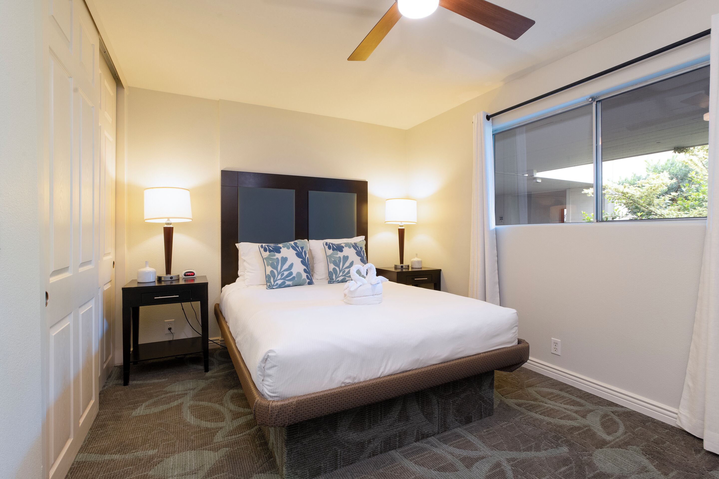 A hotel room with a large bed, two bedside tables with lamps, a ceiling fan, and a window with a view of greenery.