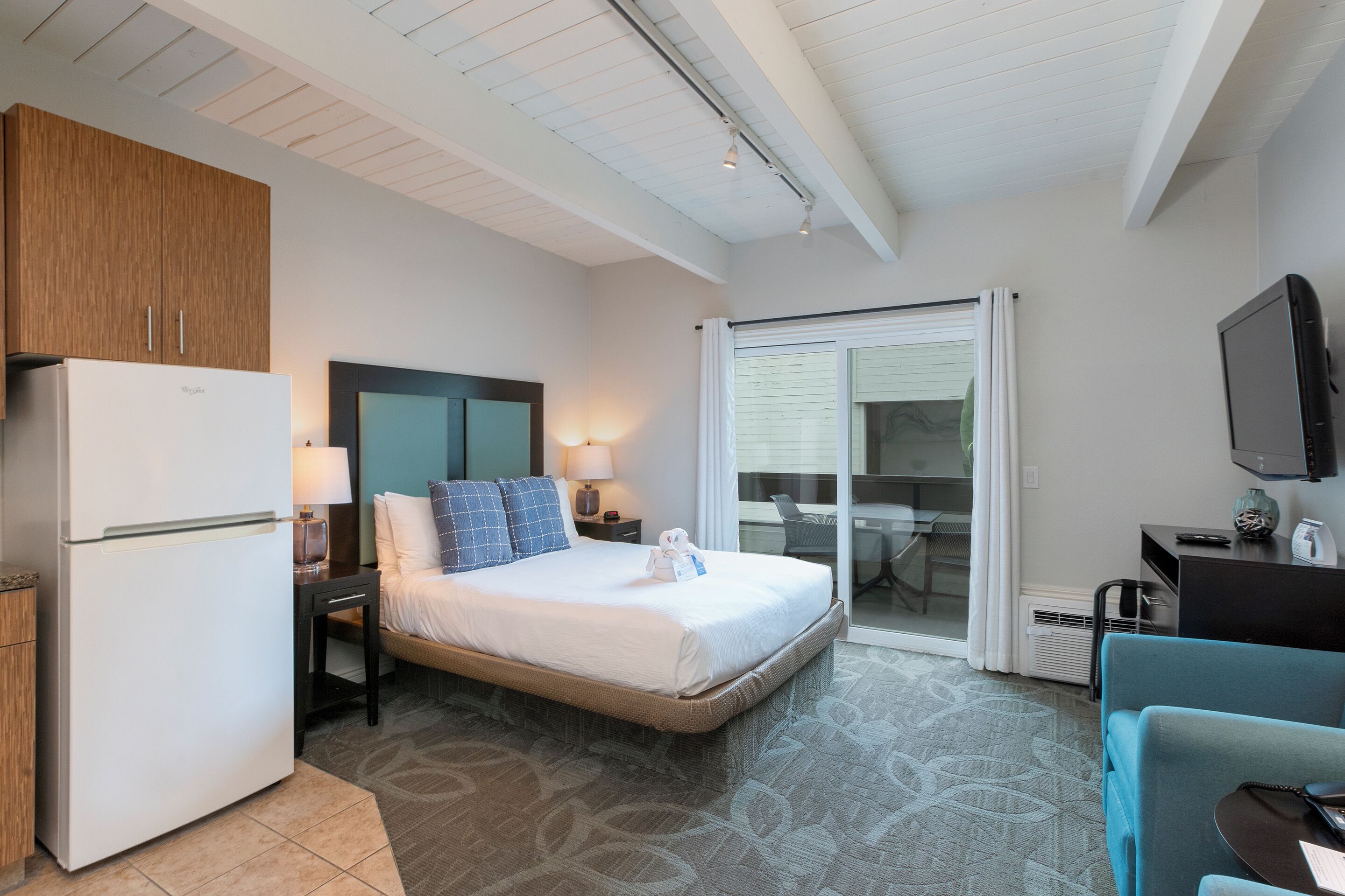 A hotel room with a bed, a refrigerator, a desk with a chair, a television, and a sliding glass door to a balcony.