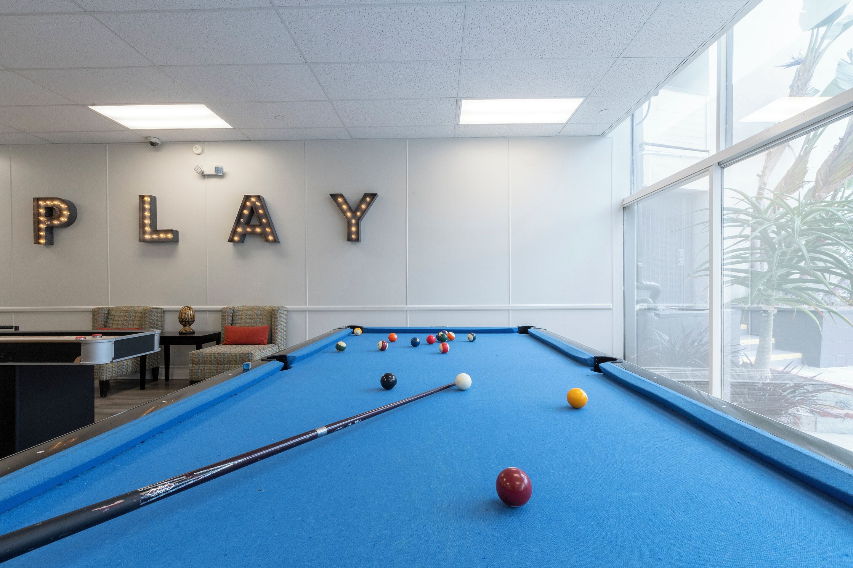 A pool table with balls, a pool table, a sofa, and a wall with illuminated letters spelling "PLAY".