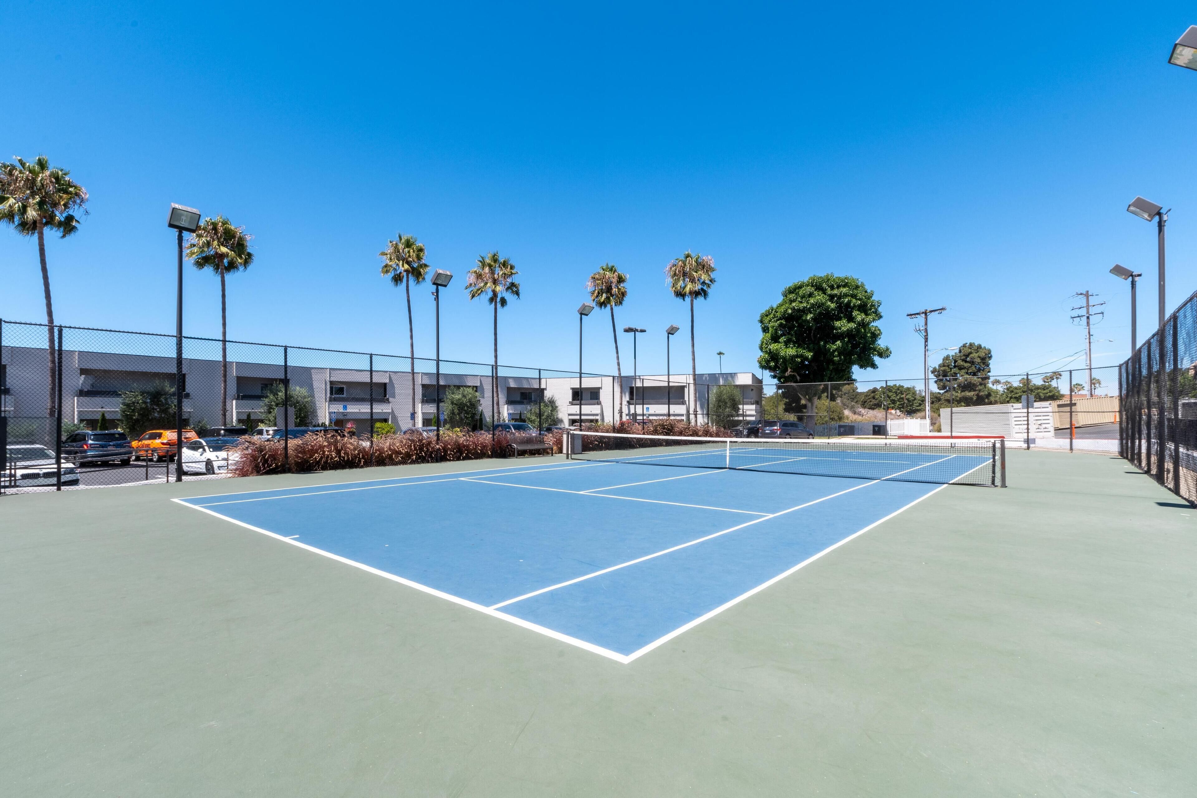 An outdoor tennis court with a net, surrounded by a fence, palm trees, and a building in the background.