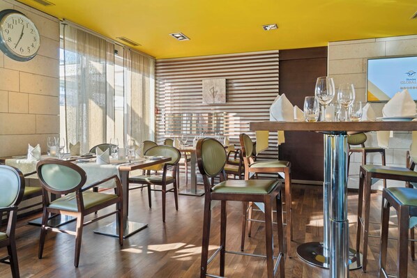 A restaurant with wooden tables and chairs, a clock on the wall, and a large window with blinds. - Hotel Conti Dietikon (Dietikon)