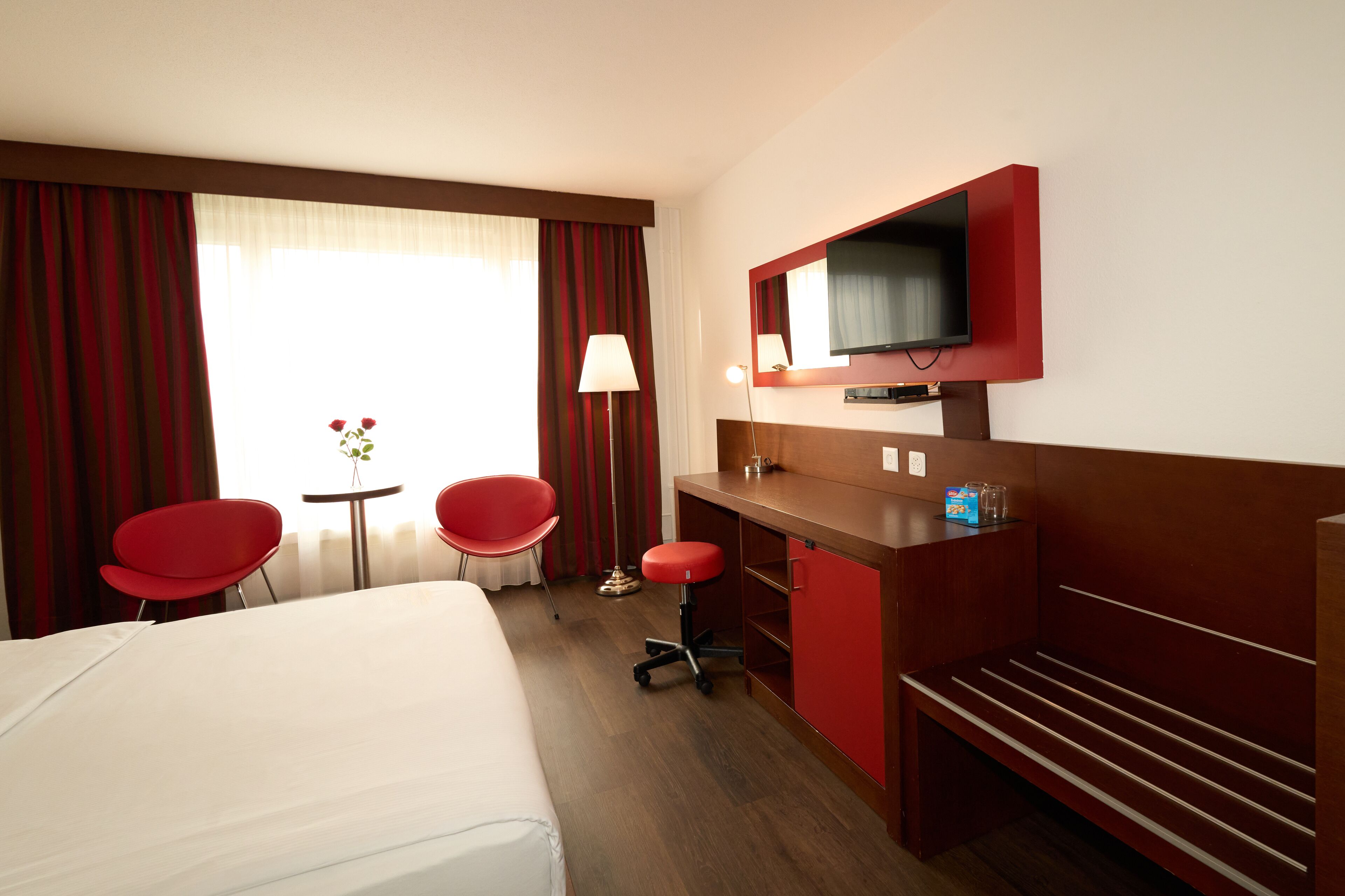 Une chambre d’hôtel avec un lit, deux fauteuils rouges, un bureau avec une télévision et une fenêtre avec des rideaux.